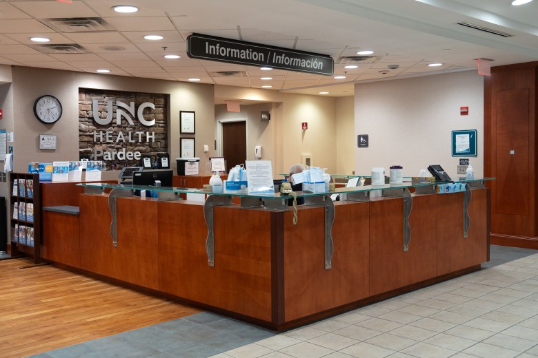 Shows the reception desk inside of Pardee Hospital in Henderson County. The large wooden desk has a glass top with a landline phone and hand sanitizer container on one corner.