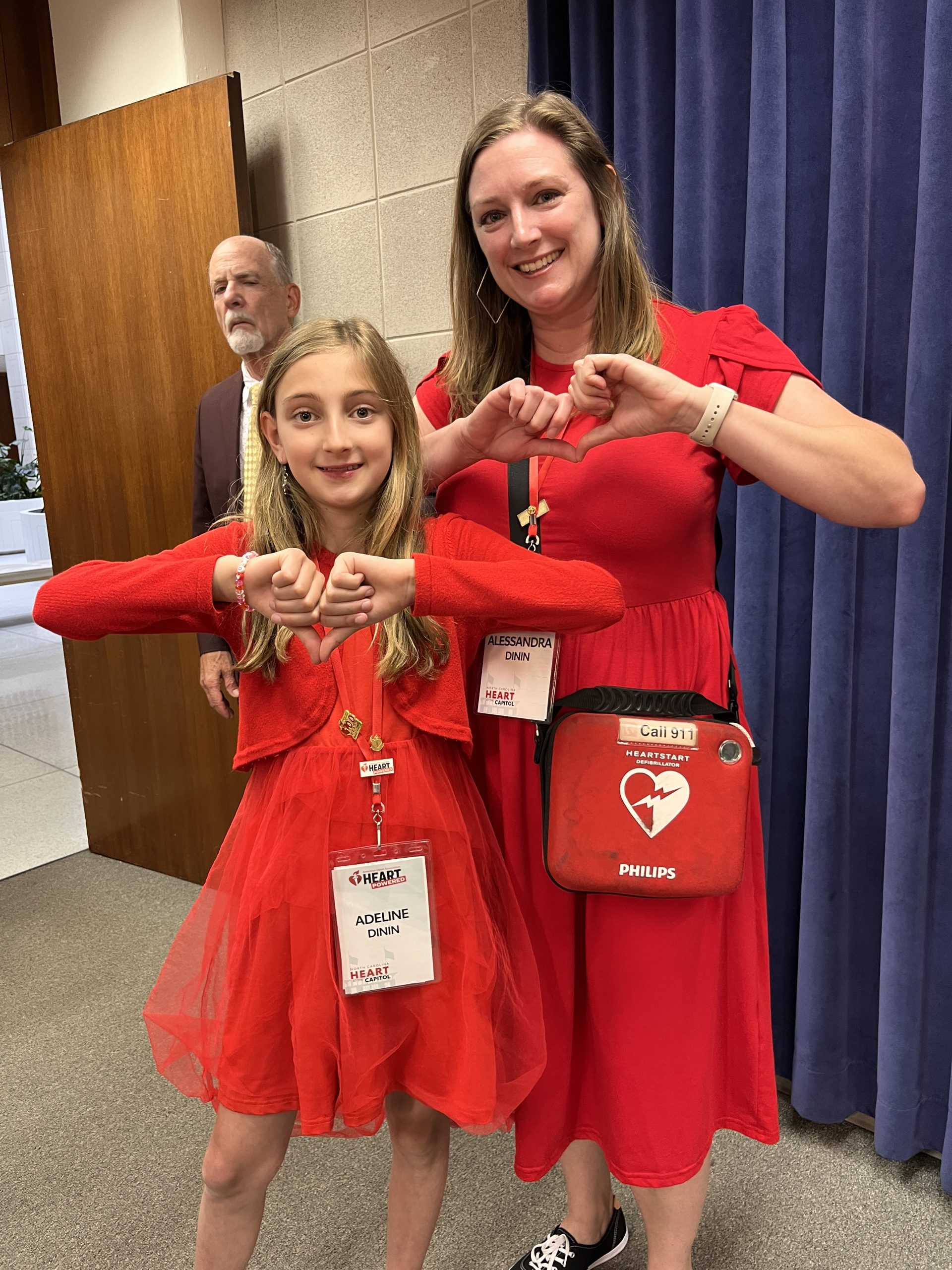 a young woman and her mother, both wearing red, make heart symbols with their hands. The woman is carrying an automatic external defibrillators in a pouch hanging from her shoulder.