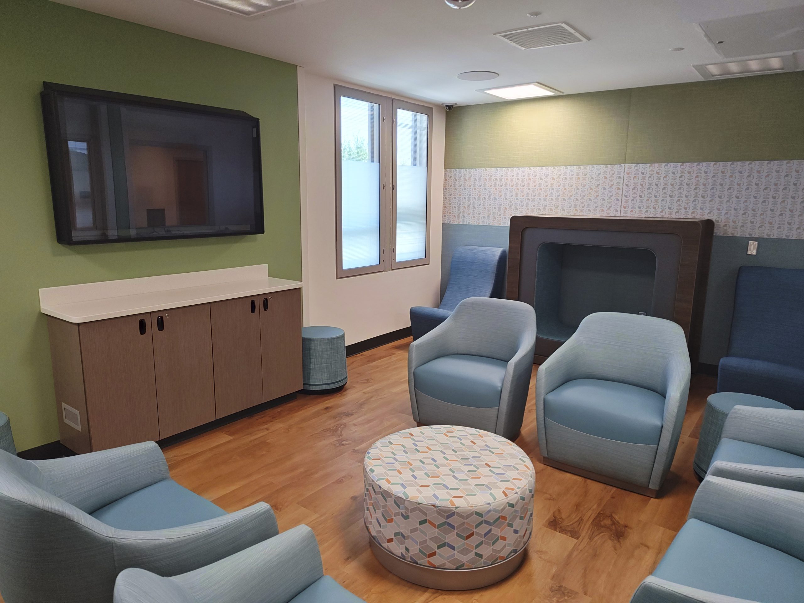 A room with green walls is filled with blue chairs. There are two big windows on one wall next to a large, wall-mounted TV screen.