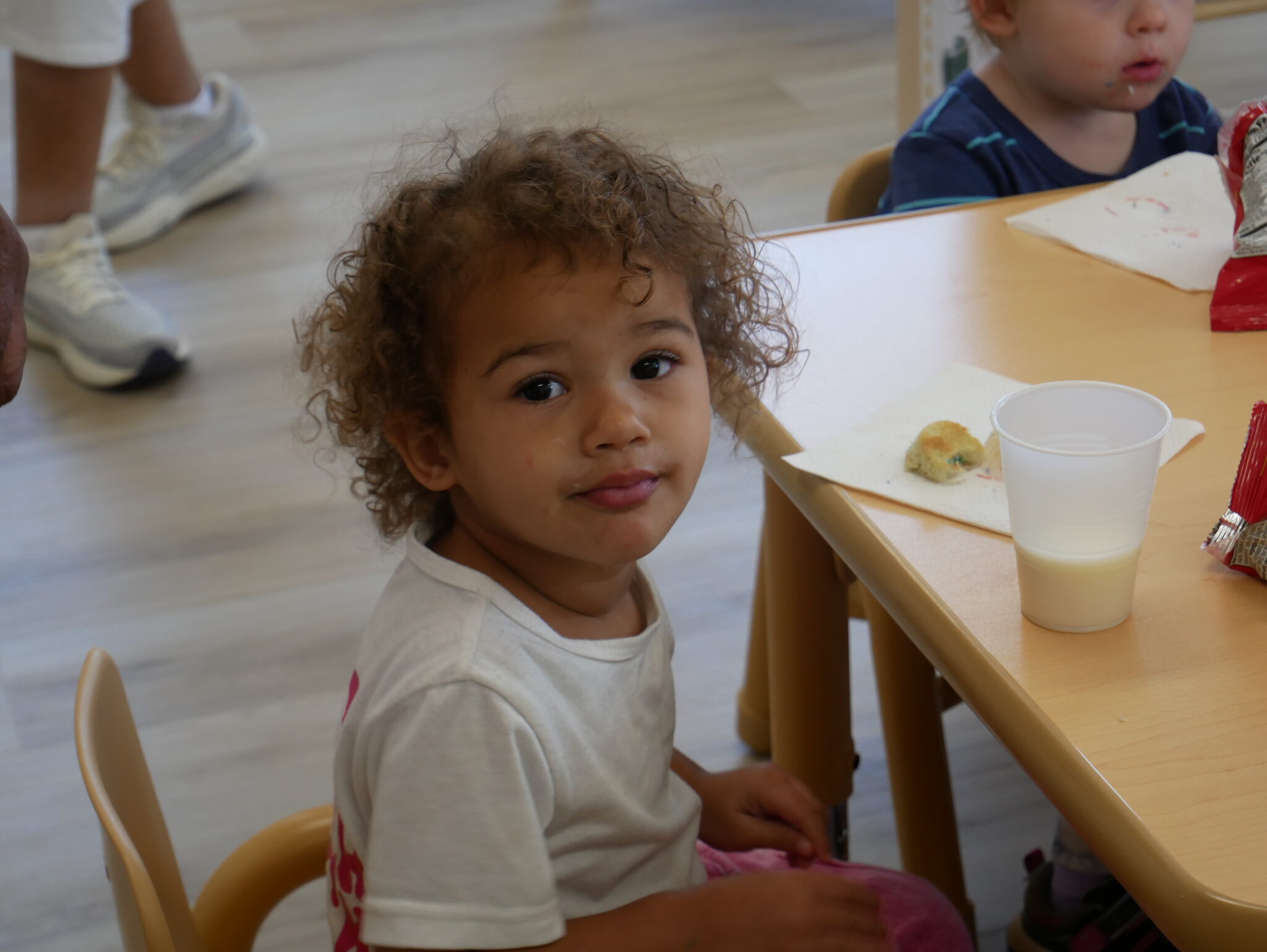 A young child sits at a table in a childcare classroom, smiling at the camera during an activity.