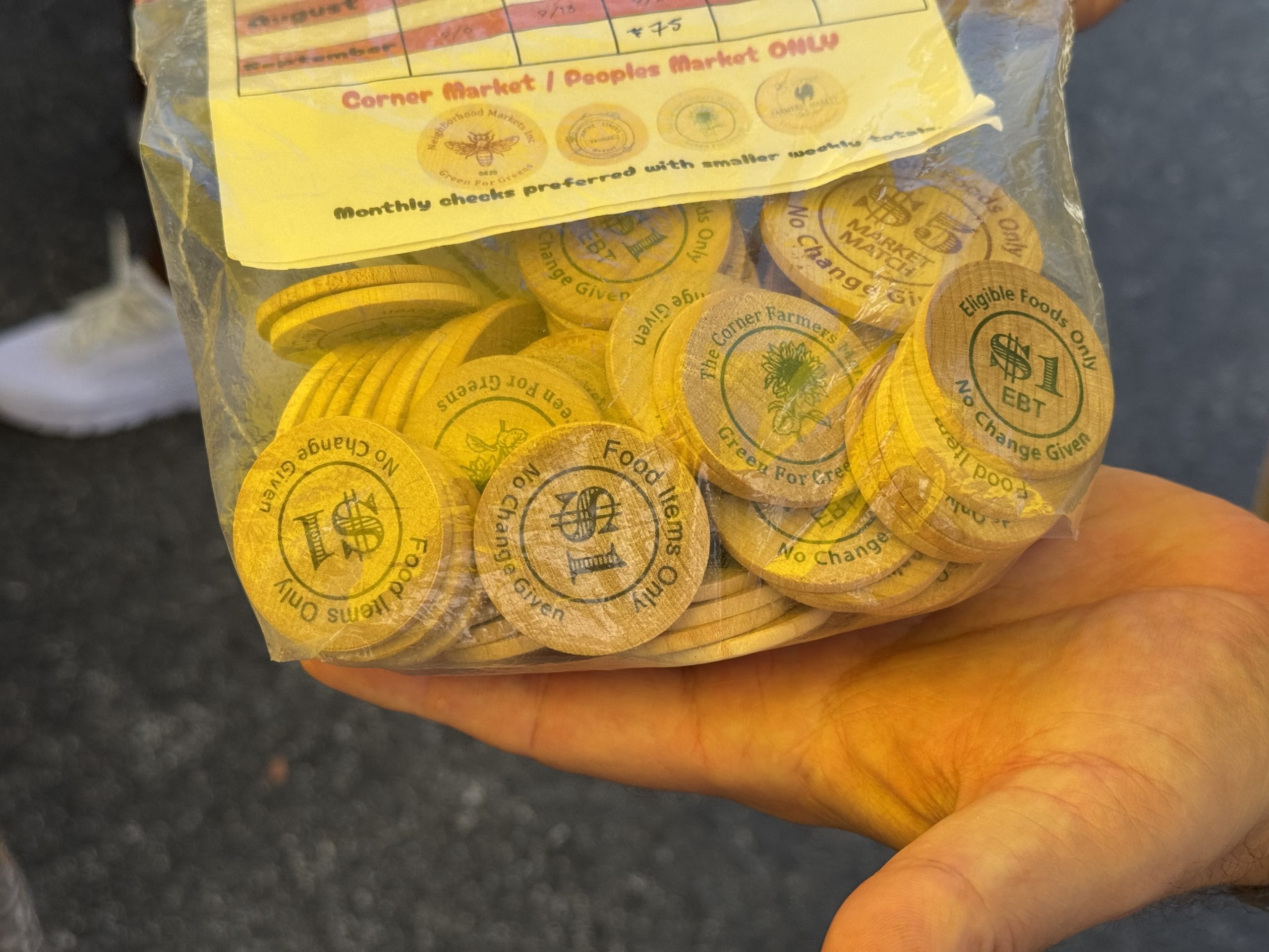 A closeup of a hand holding a plastic bag filled with wooden market tokens valued at one and five dollars.