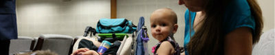 shows a young woman with an infant on her lap and another young child in a wheelchair with her. BOth childrem have the genetic disease SMA.