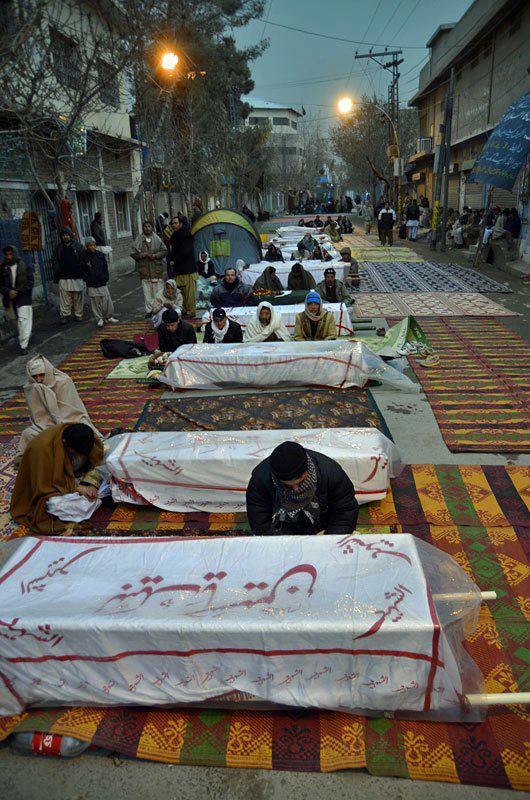 Shows a line of coffins in the road each covered by a cloth, with people sitting near them, huddled against the cold.