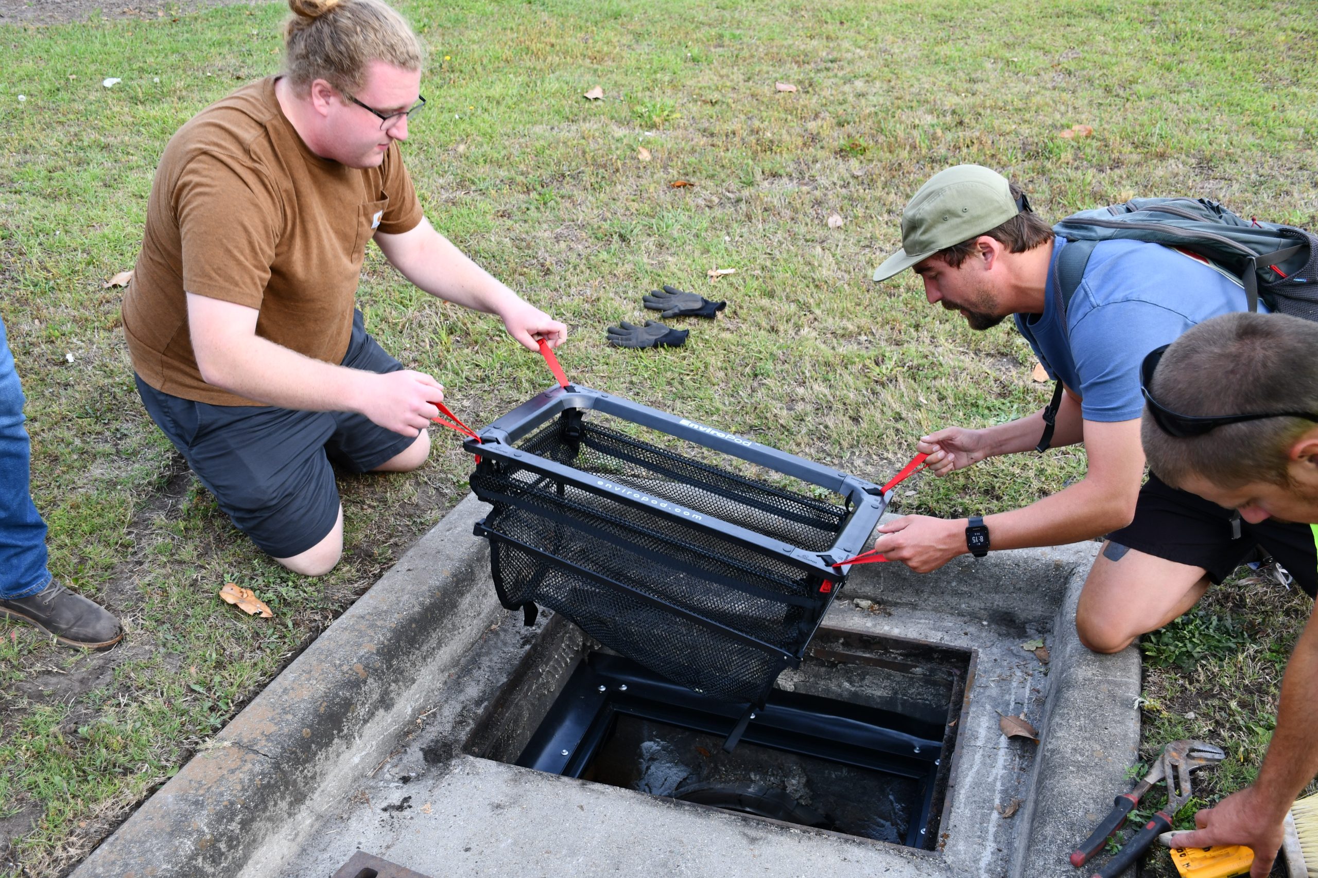 Shows two men kneeling as they place a black basket with mesh netting, called a "Litta Trap," into a concrete catch basin to trap small-sized debris before it enters local waterways