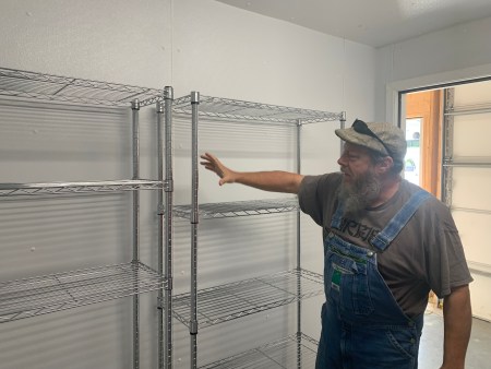 A man wearing a grey t-shirk and blue overalls and cap with sunglasses resting on it, points at a row of metal shelves located in a room with white walls.
