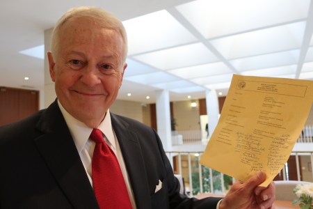 white man in a suit jacket and a red tie holds up a paper with printing and handwriting on it