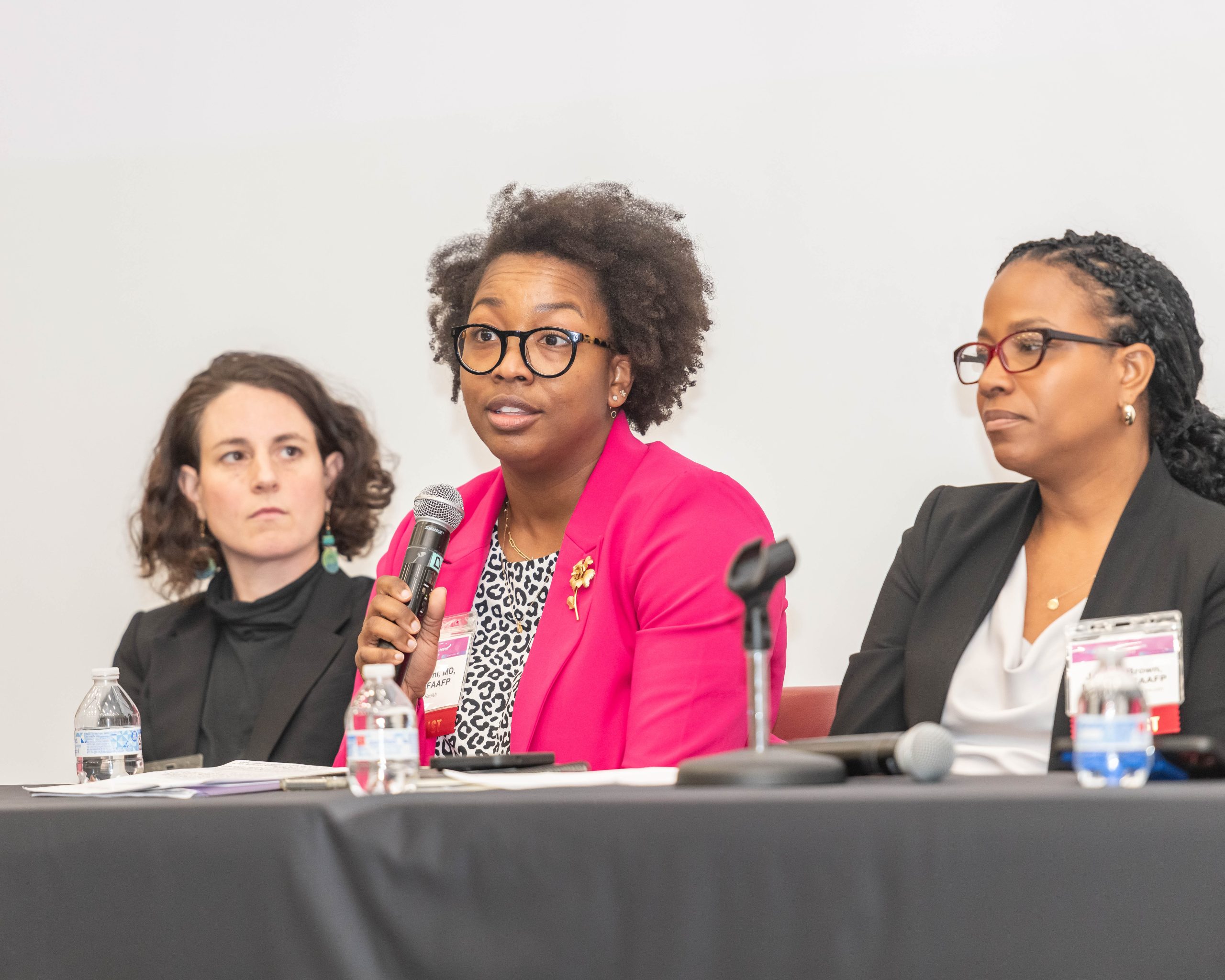 A Black woman wearing a bright pink blazer holds a microphone and speaks on a panel at a Black maternal health conference