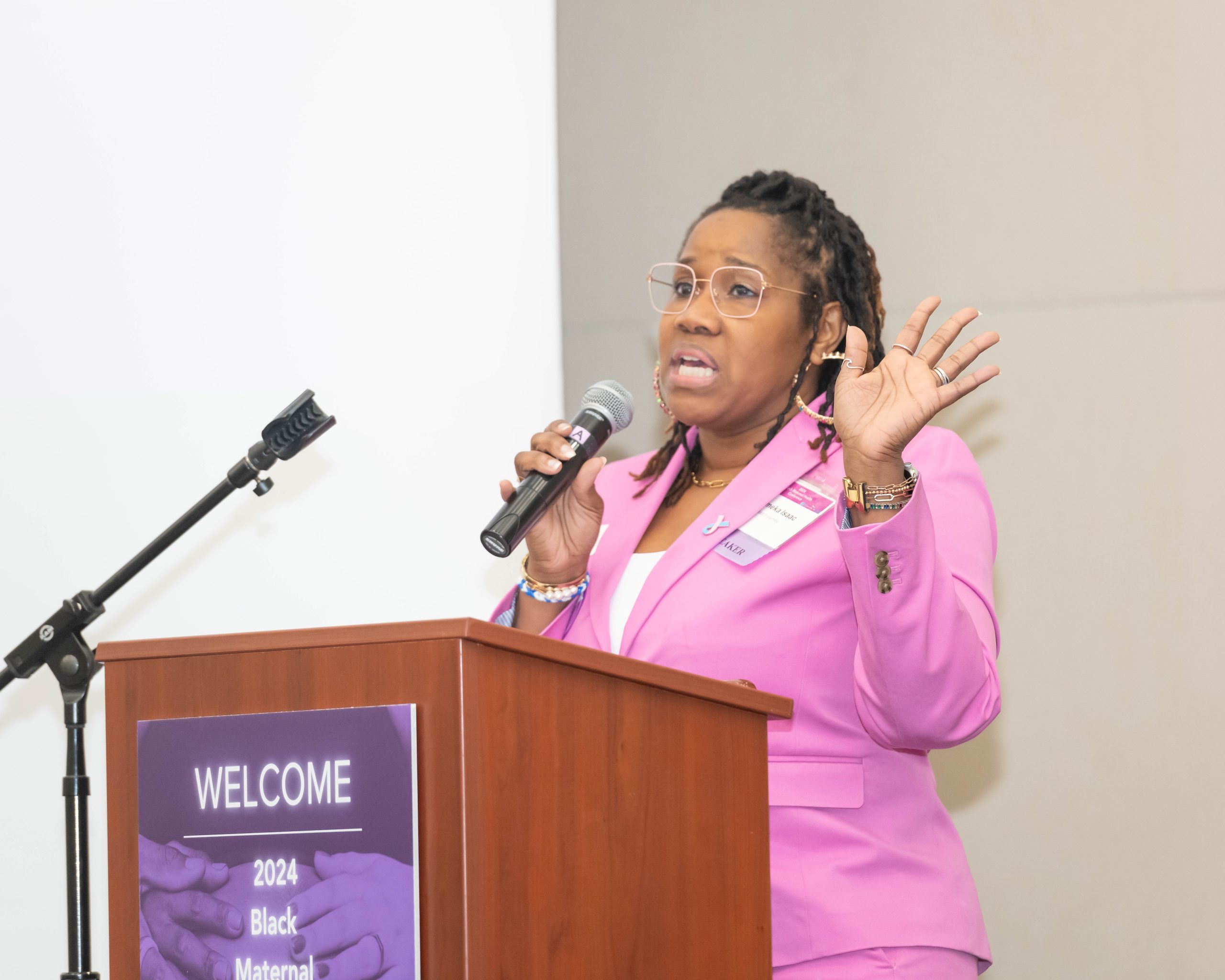 A Black woman wearing a bright pink suit shares her personal experience with pregnancy complications and the loss of her son at a conference on Black maternal health