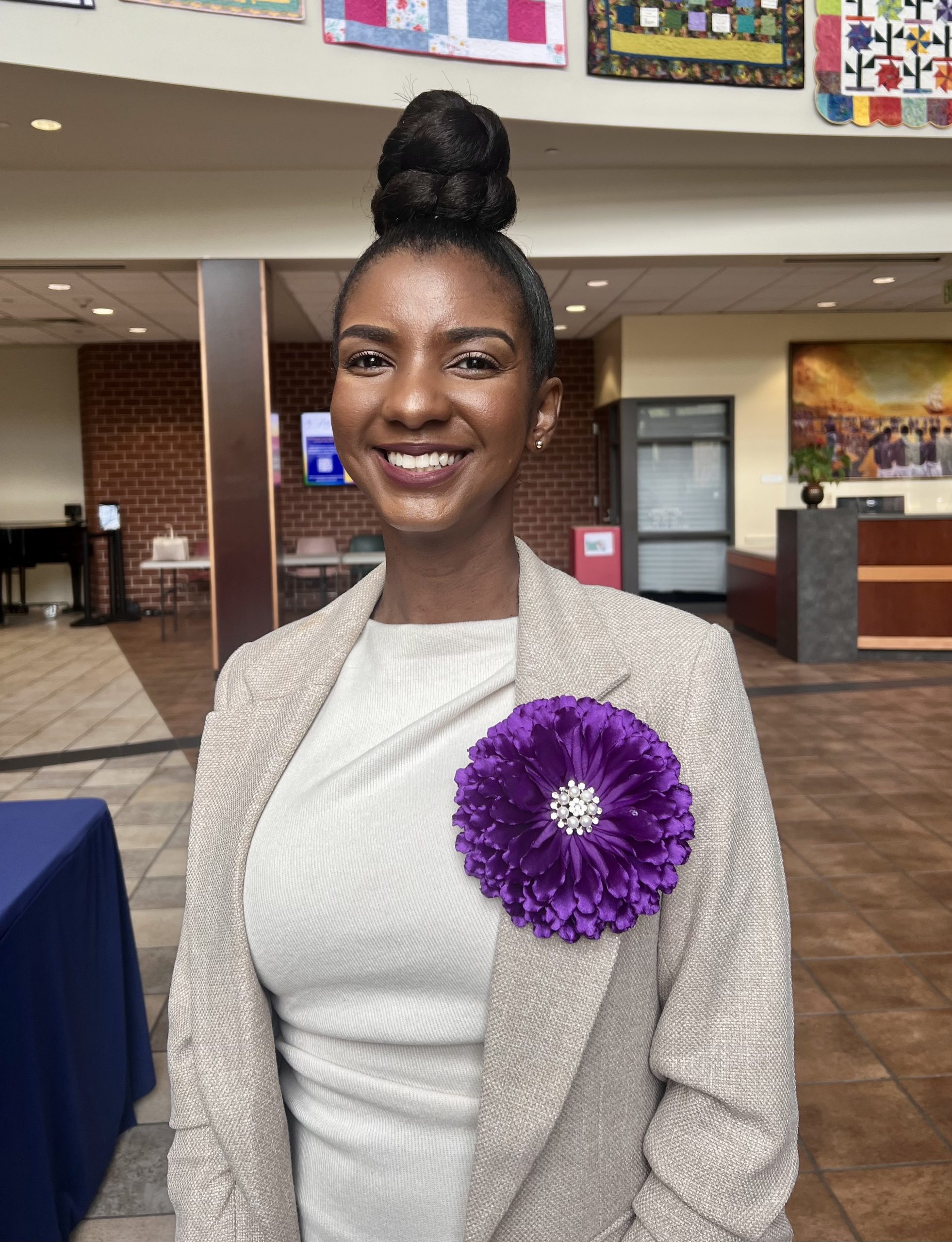 A Black woman in a tan dress with a purple flower on the lapel smiles for a photo