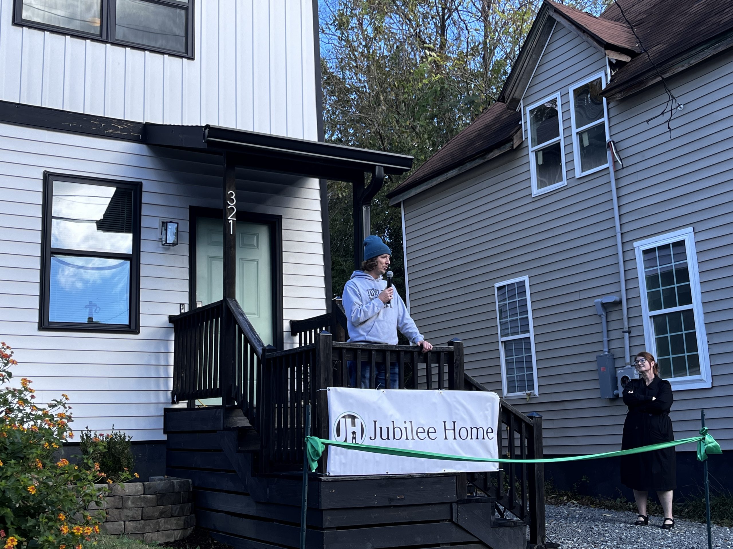 A man in a hat stands on the stairs of a house holding a microphone talking about the opening of a new women's house offering housing to formerly incarcerated women as they reenter the community