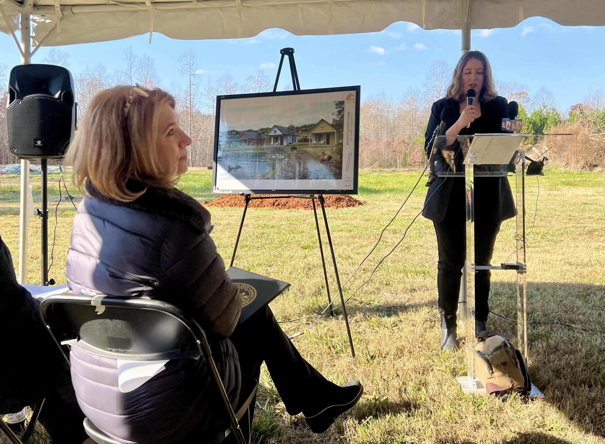 A woman stands at a podium talking about the construction of tiny homes for formerly incarcerated women. A lady seated in the audience is shown looking at an image rendering of the housing.