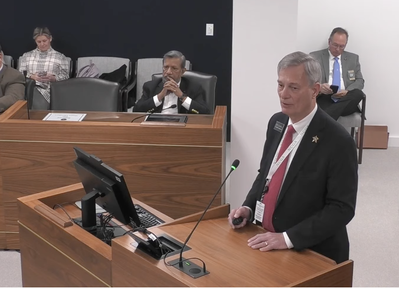 A man in a suit stands at a podium talking to lawmakers about Iryna's Law