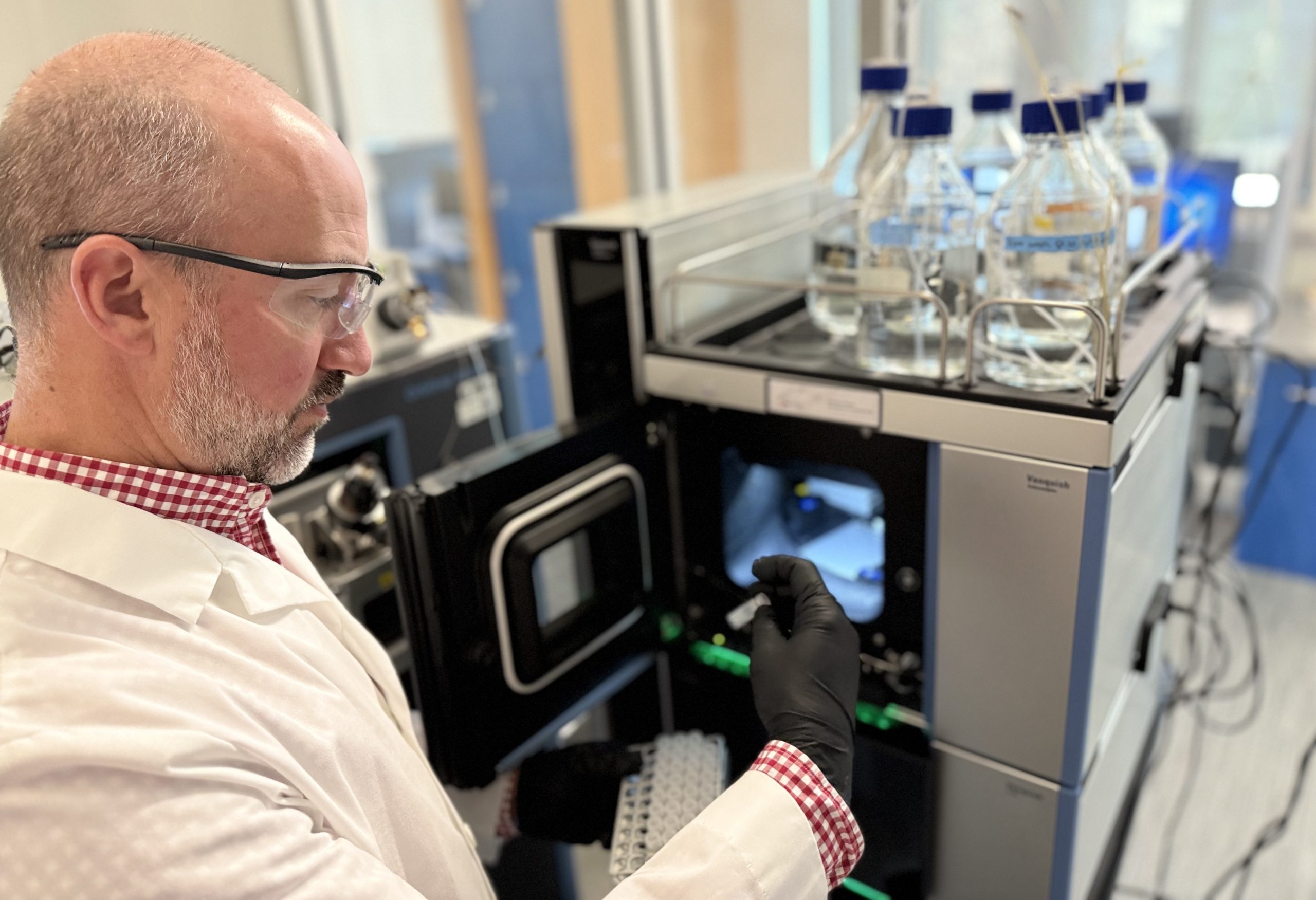 A man wearing safety glasses, black gloves and a white lab coat examines a small test tube he has just removed from an analytical laboratory instrument.