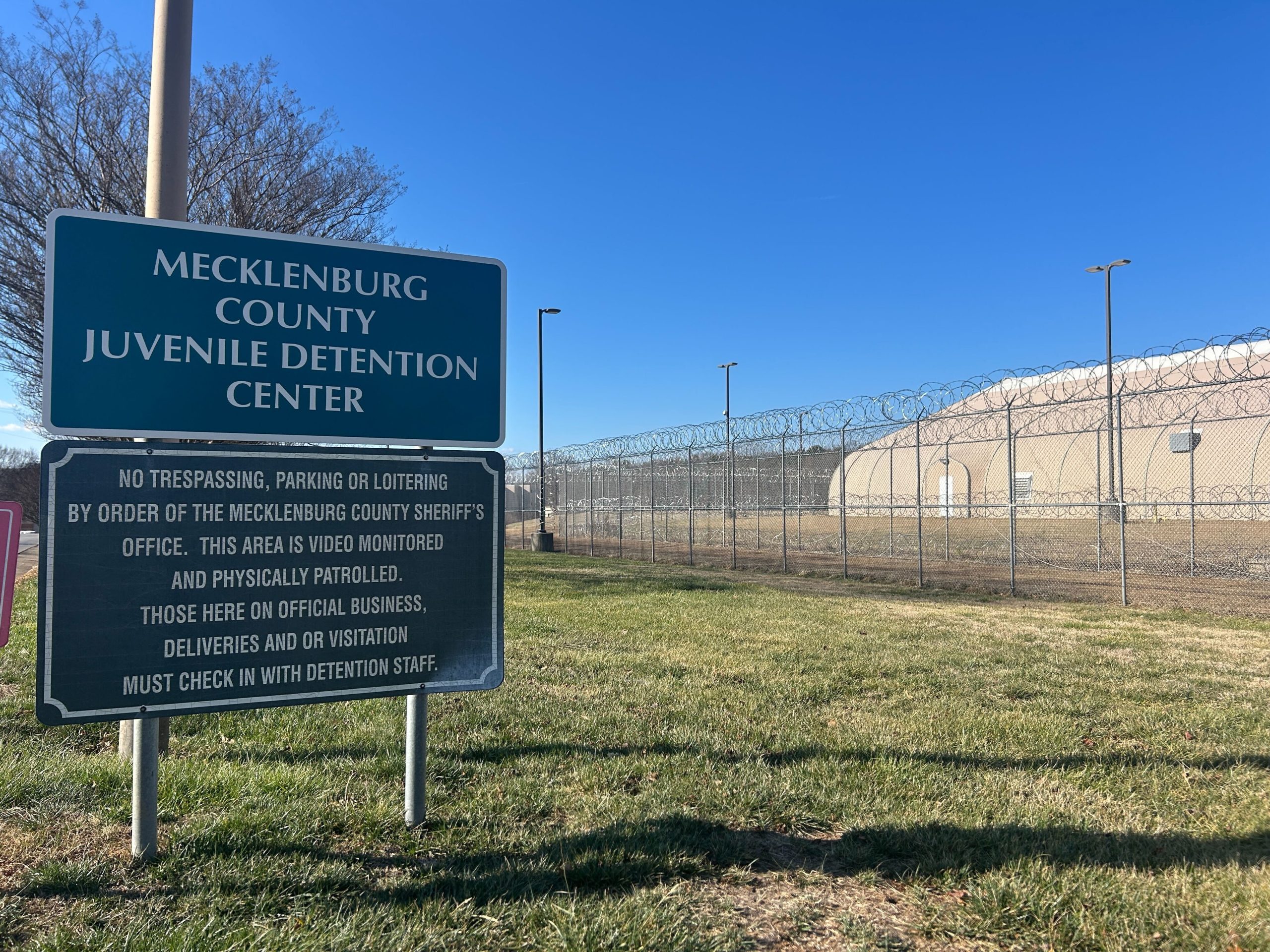 A blue sign marking the Mecklenburg County Juvenile Detention Center. The center is in the background lined with barbed wire fence