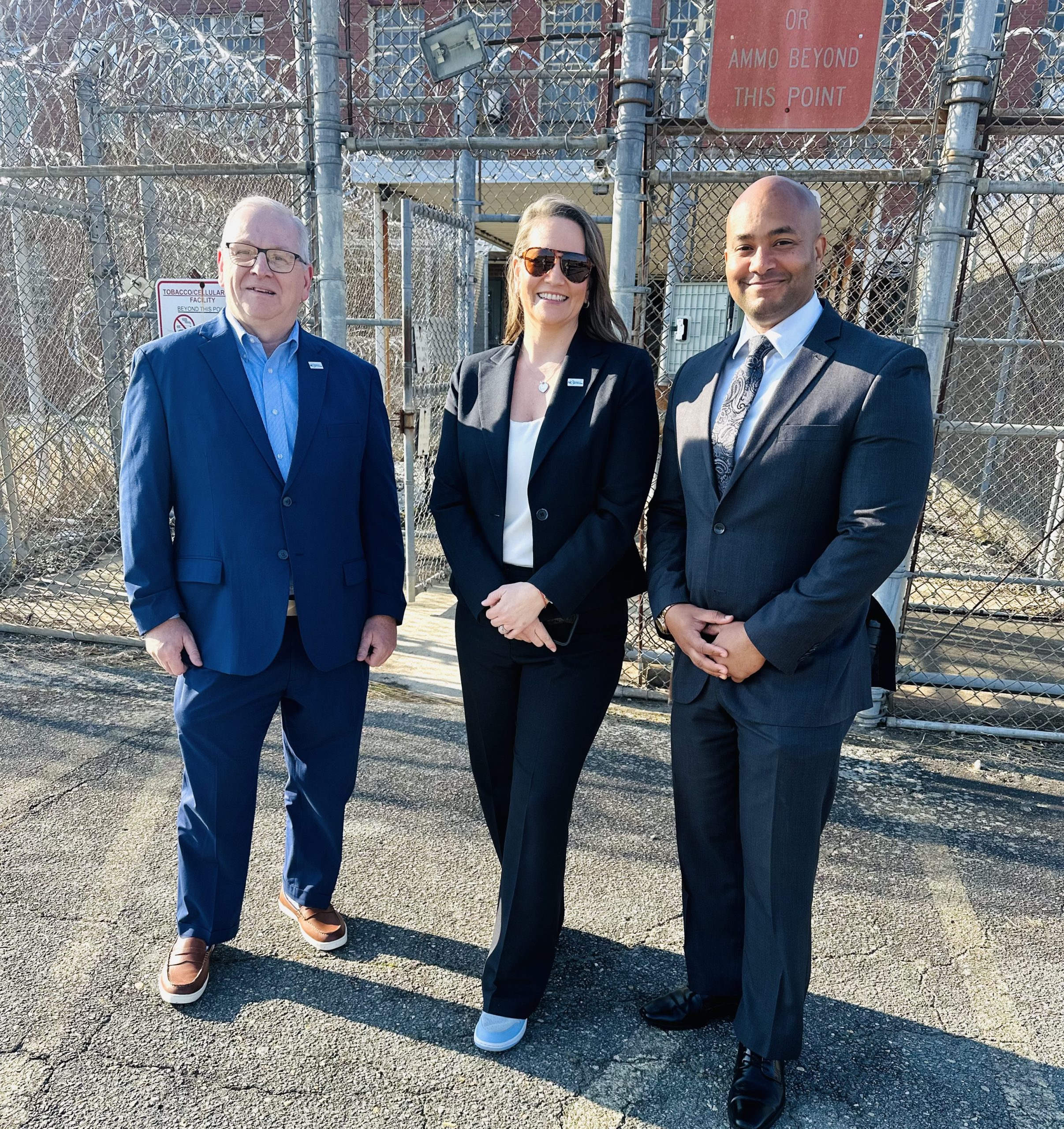 Three people stand in front of a prison barbed wire fence