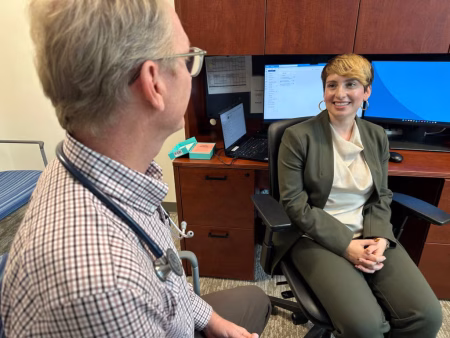 White older man in the foreground with a stethoscope around his neck and glasses looking at a younger woman in business attire with short hair and computer monitors behind her.