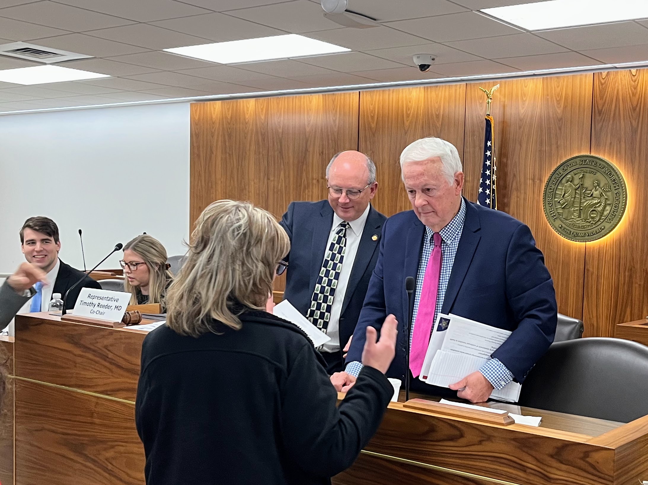 two older white men behind a podium talk with an older woman whose back is to the camera after a meeting of the House Select Committee on Involuntary Commitment and Public Safety.
