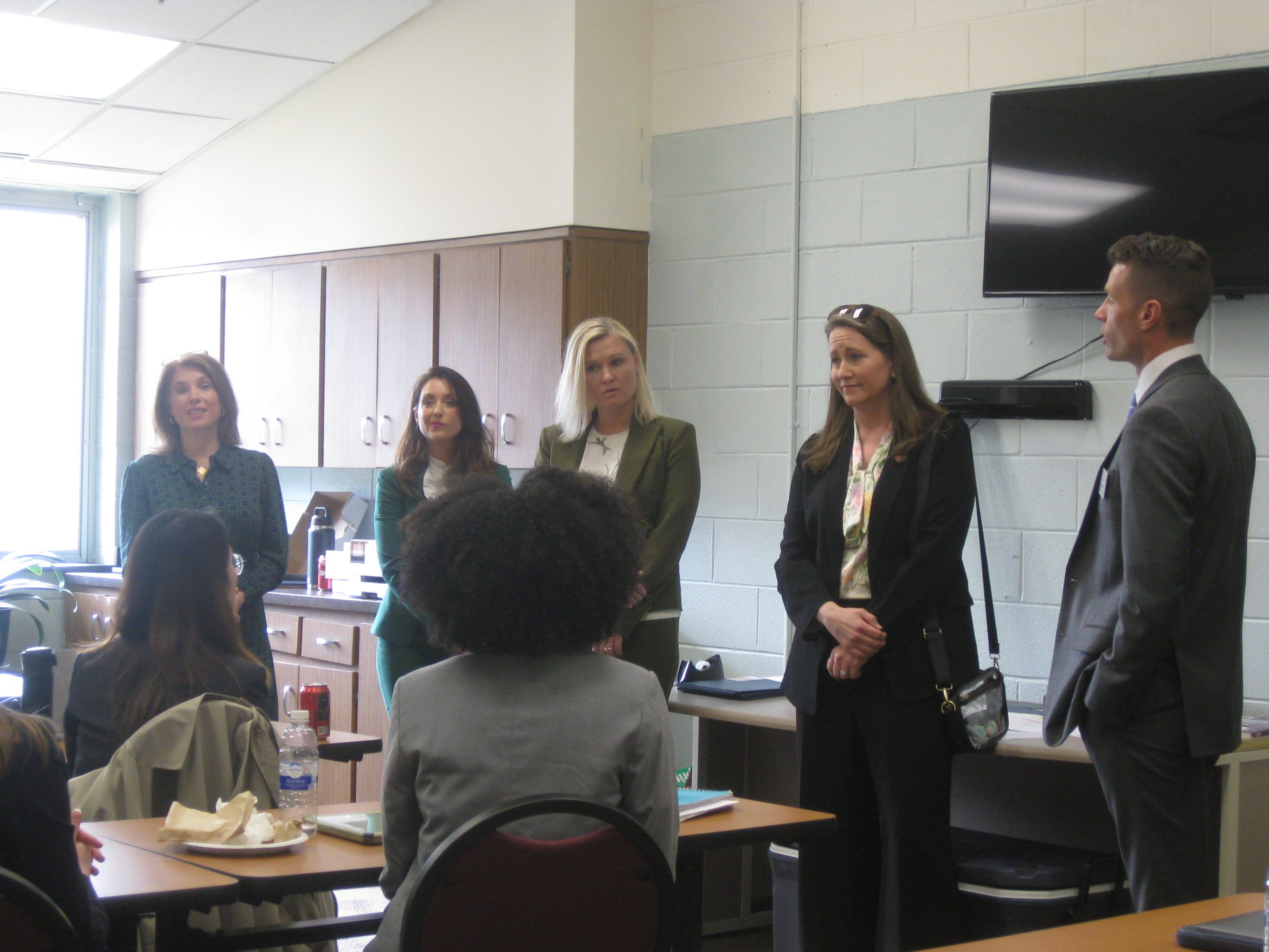 Four women and a man stand in a room in front of law students assisting with a driver's license restoration clinic at a prison.