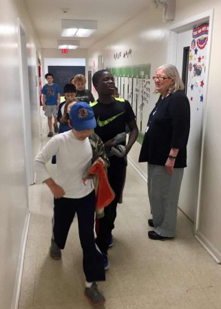 older white woman stands in a school hallways greeting students walking by in a line