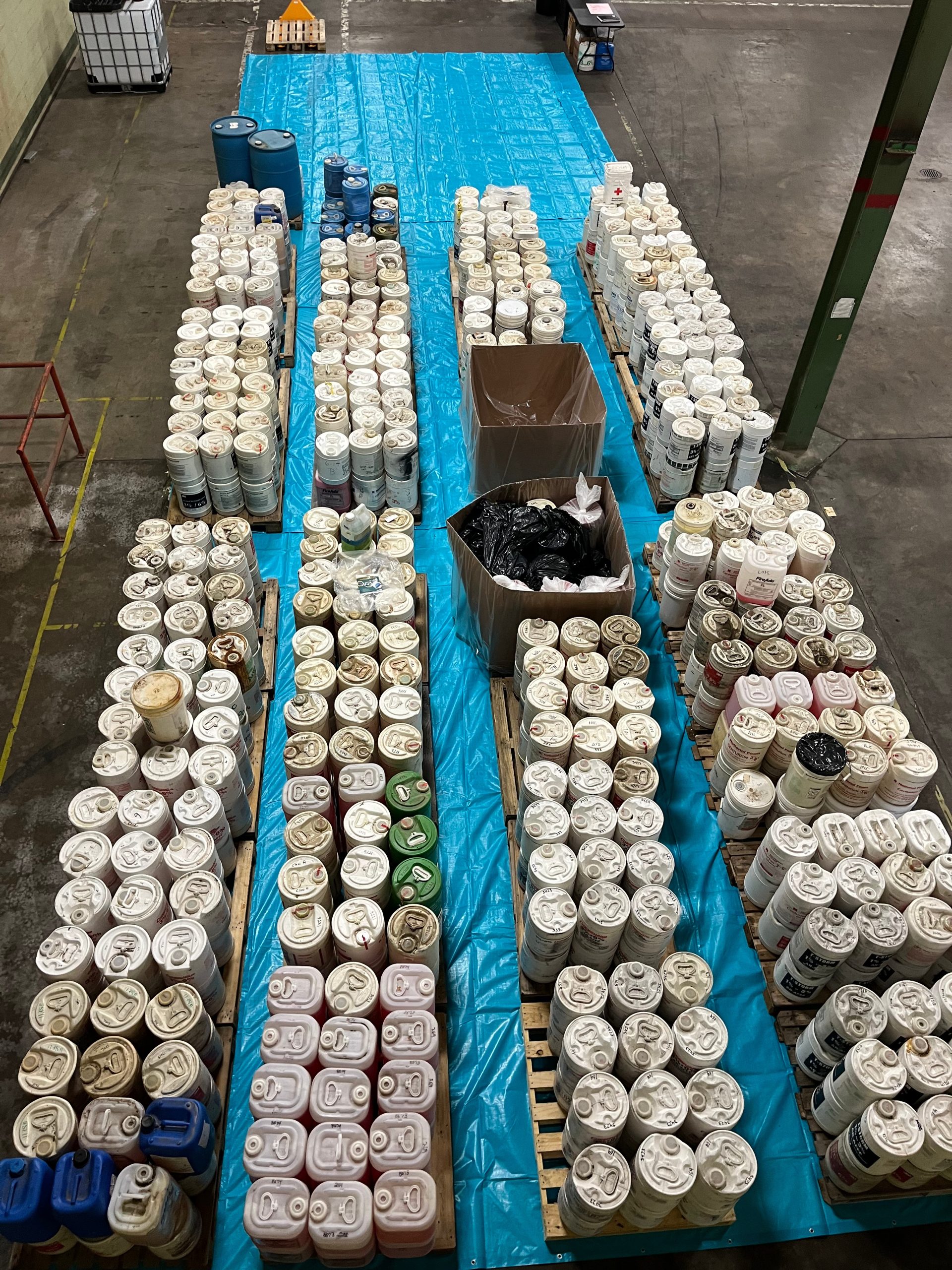 n overhead view inside a warehouse, showing long rows of wooden pallets covered by a bright blue plastic tarp and stacked tightly with hundreds of white plastic buckets and a few colored jugs, likely containers of chemicals or paint, with two large cardboard boxes of loose items in the middle and industrial walls and floor visible around the setup.