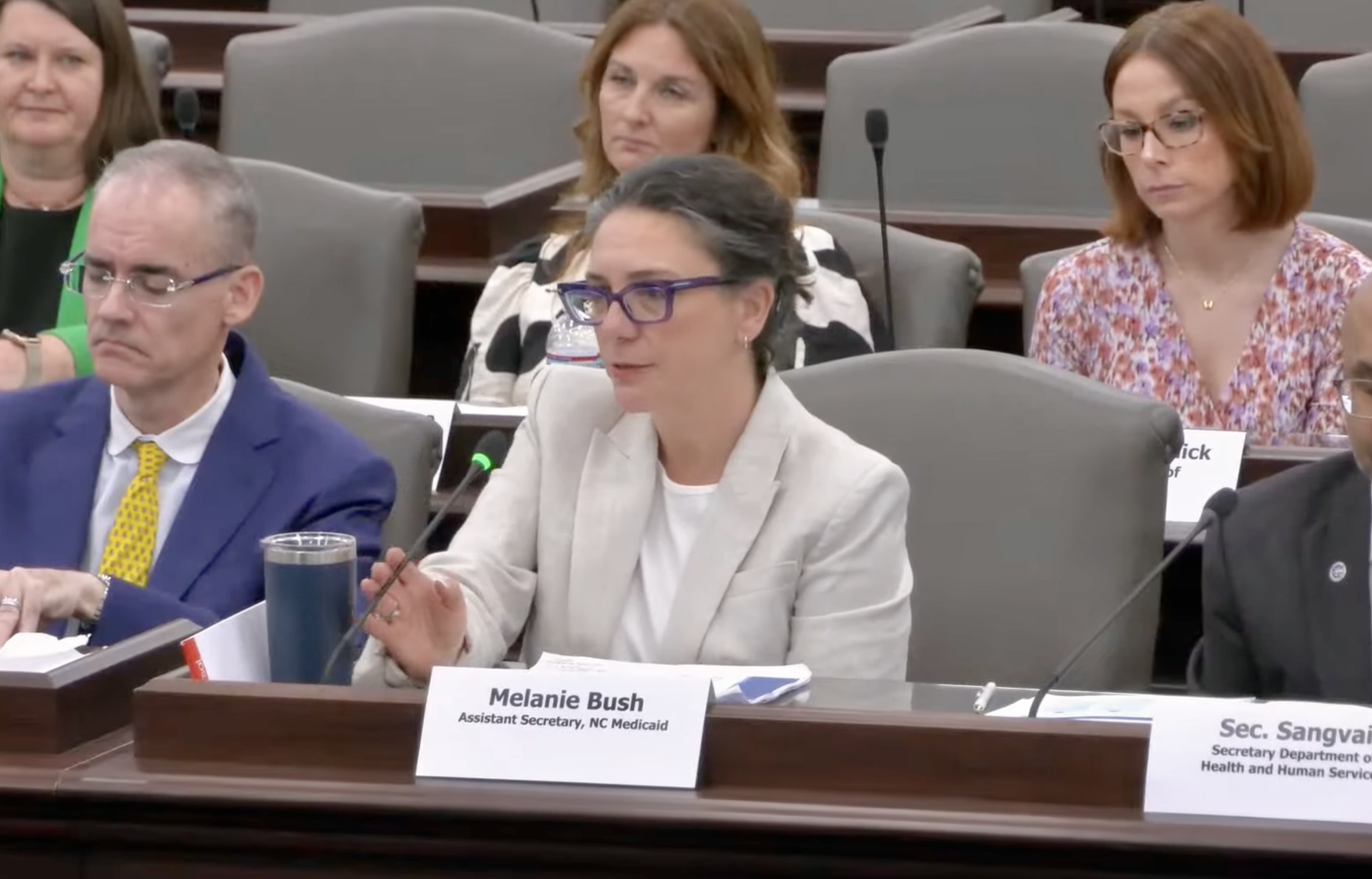 A white woman with brown hair wearing purple glasses and a light-colored suit jacket speaks into a microphone. The woman is Melanie Bush, the interim deputy secretary of NC Medicaid, who is addressing lawmakers.