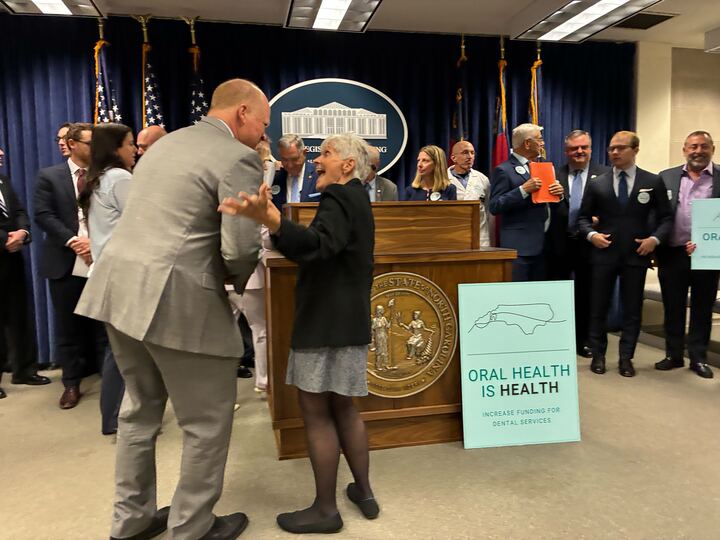 A man in a brown suit leans down to talk with a white haired woman in a black blazer.