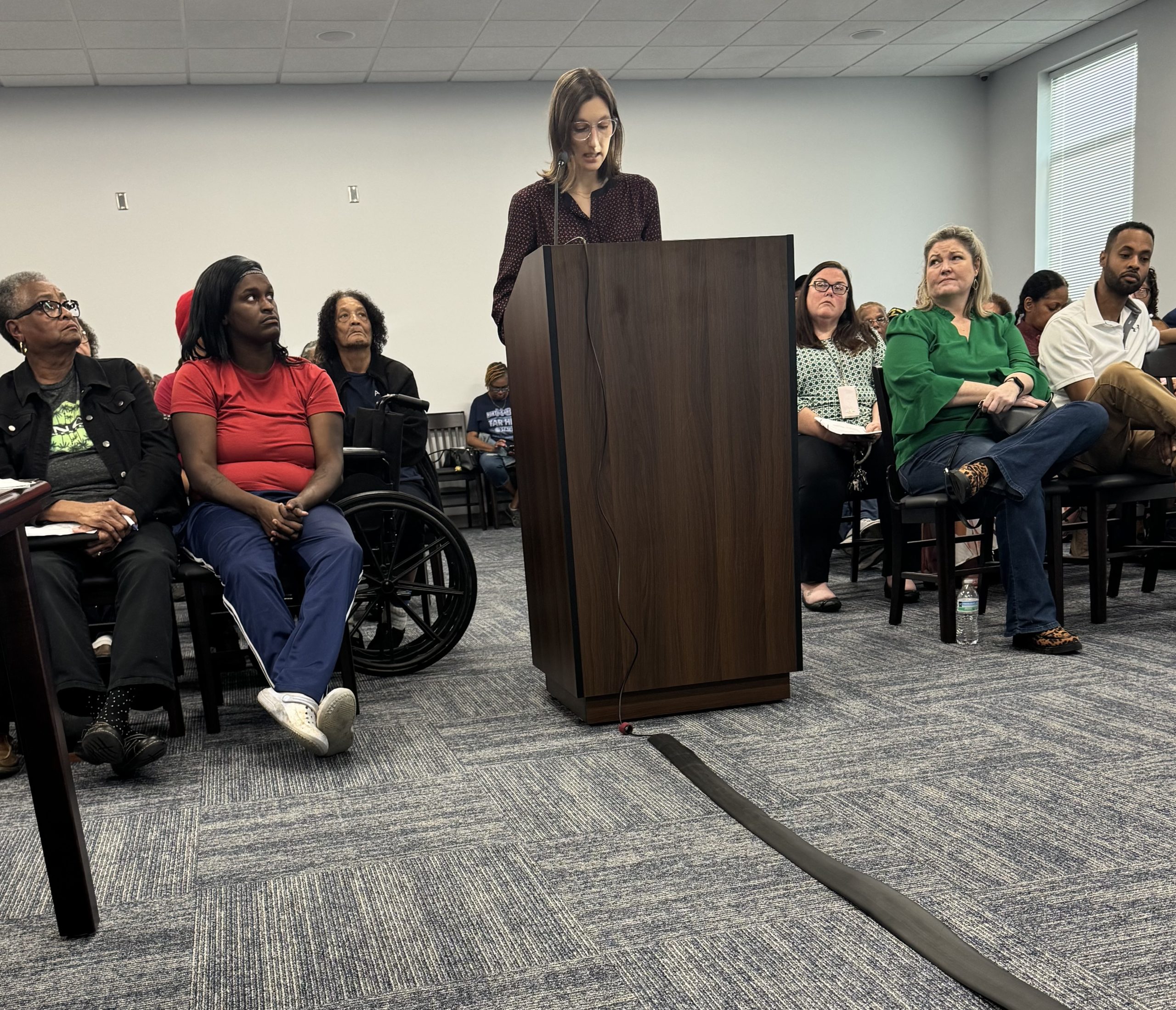 A woman stands at a podiums at a packed Northampton County commissioners meeting, urging officials to use a data center moratorium to study impacts and write rules that are fair, transparent and aligned with community values. Some audience members look at the speaker as she speaks.