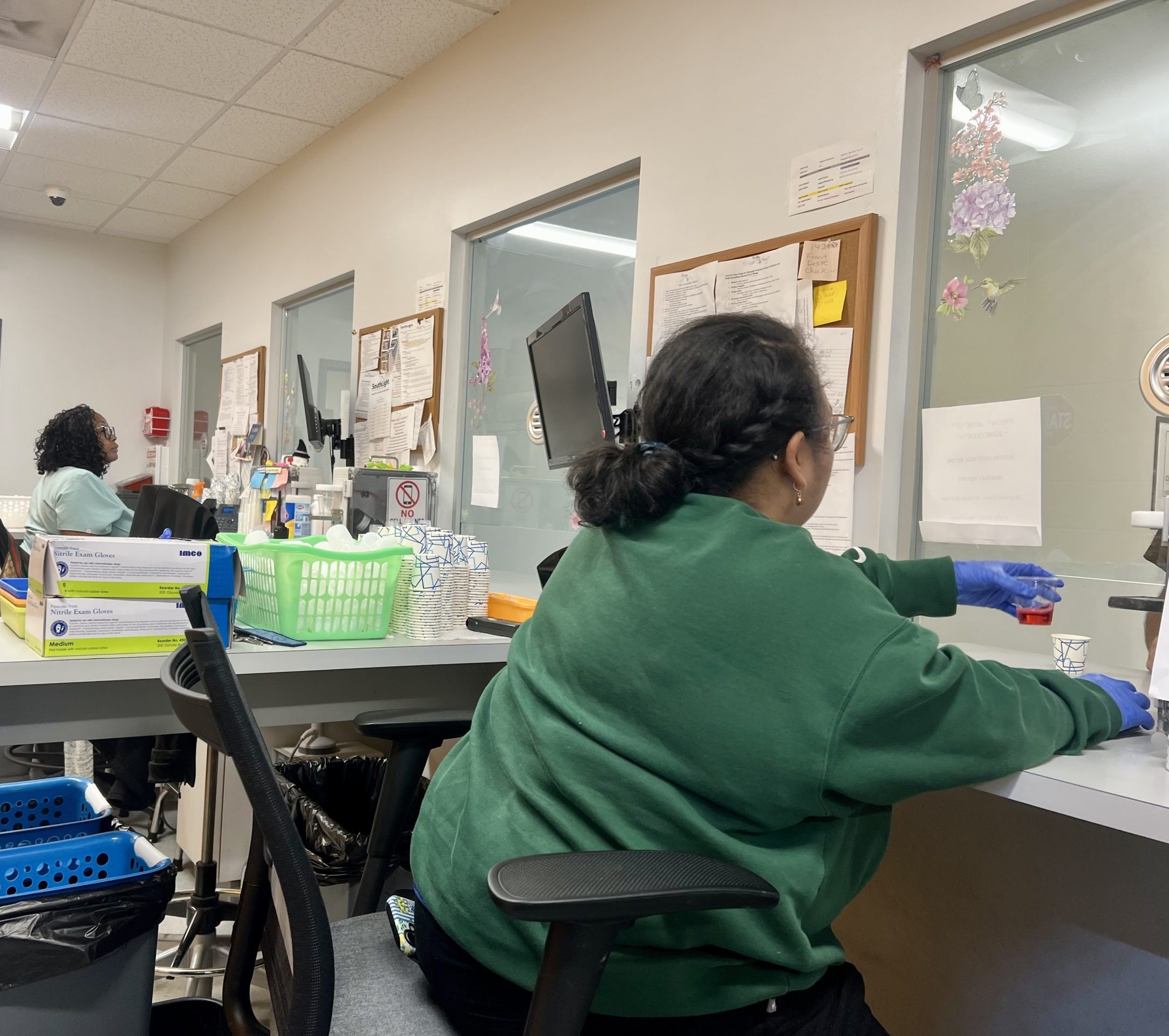 A nurse sits behind a dosing window at an opioid treatment program providing methadone to a patient