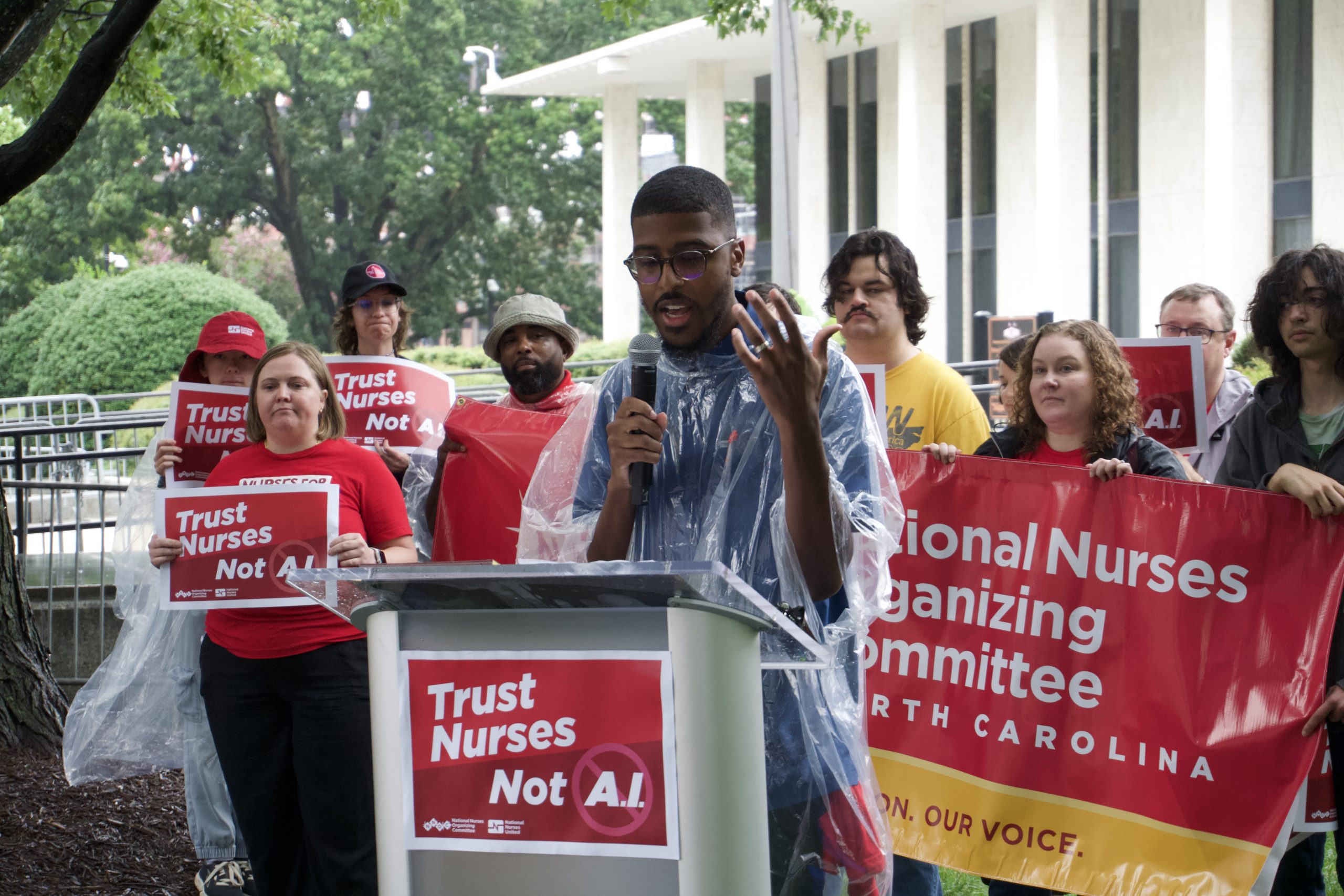Ajamu Dillahunt-Holloway speaks at a podium outside the North Carolina General Assembly as rally participants hold signs reading “Trust Nurses Not A.I.” and a National Nurses Organizing Committee banner.
