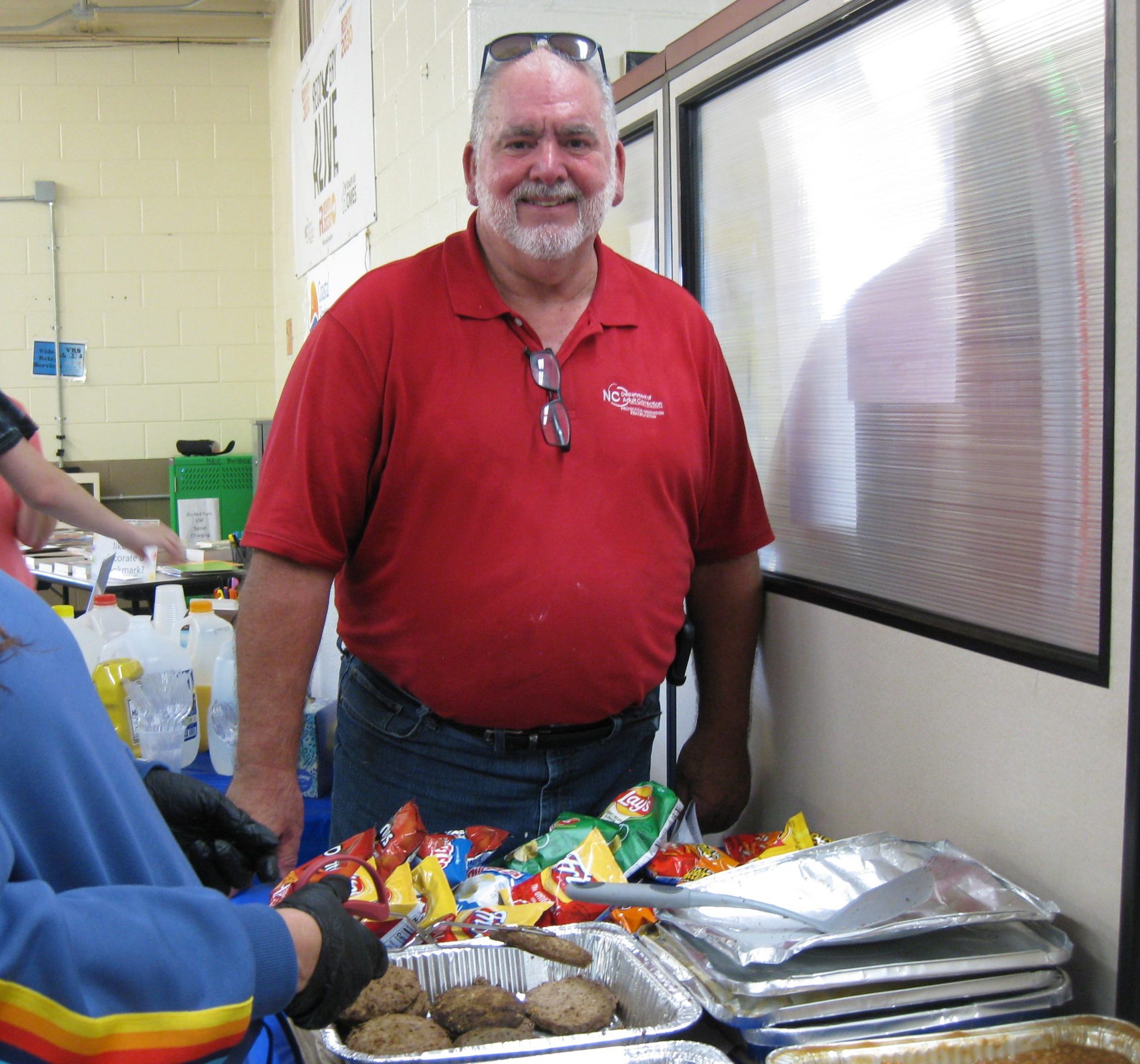 A man in a red shirt stands in front of a tray of hamburgers and chips