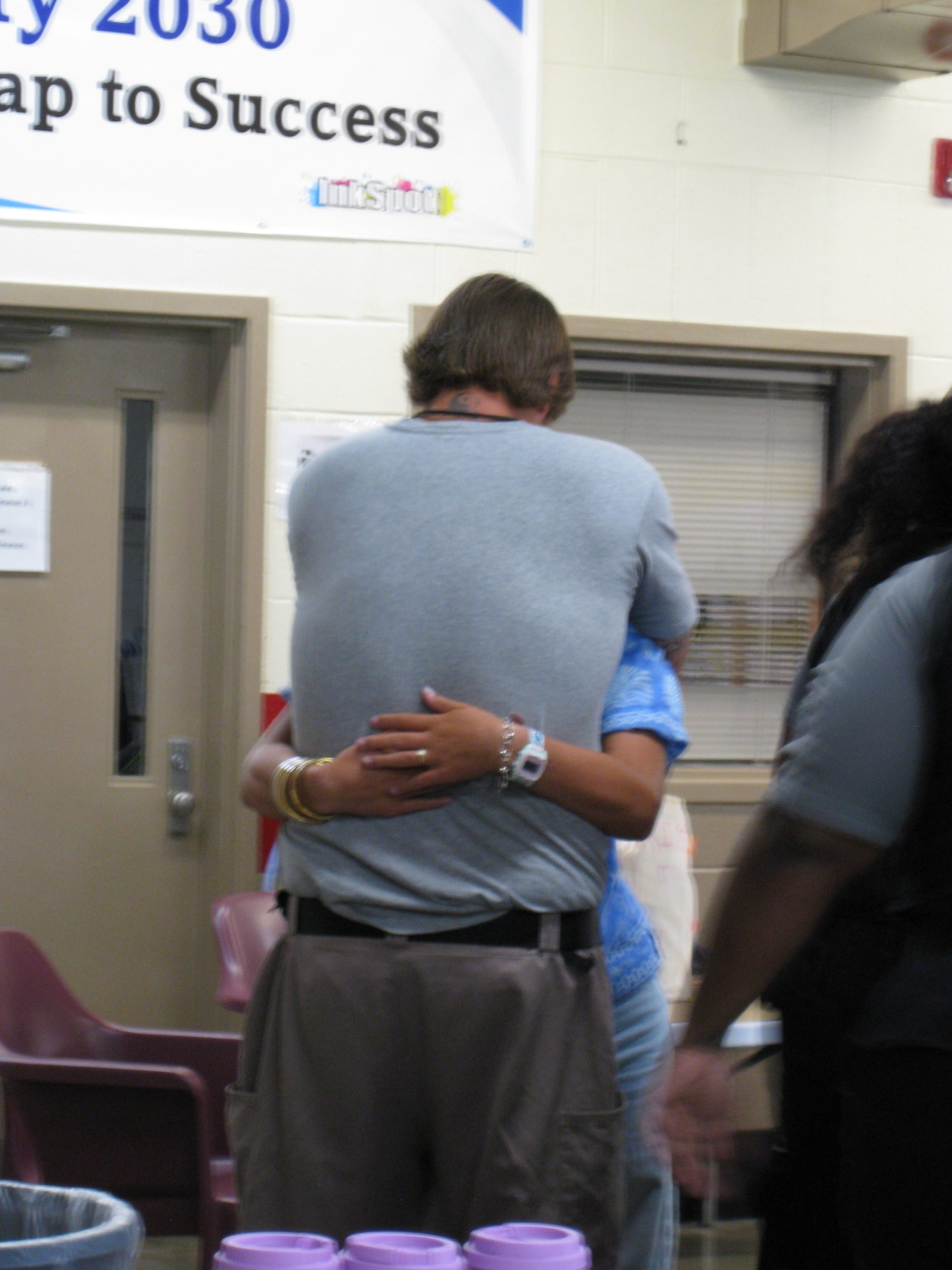 An incarcerated man in a gray shirt and brown pants tightly hugs his teenage daughter who has her hands around his waist