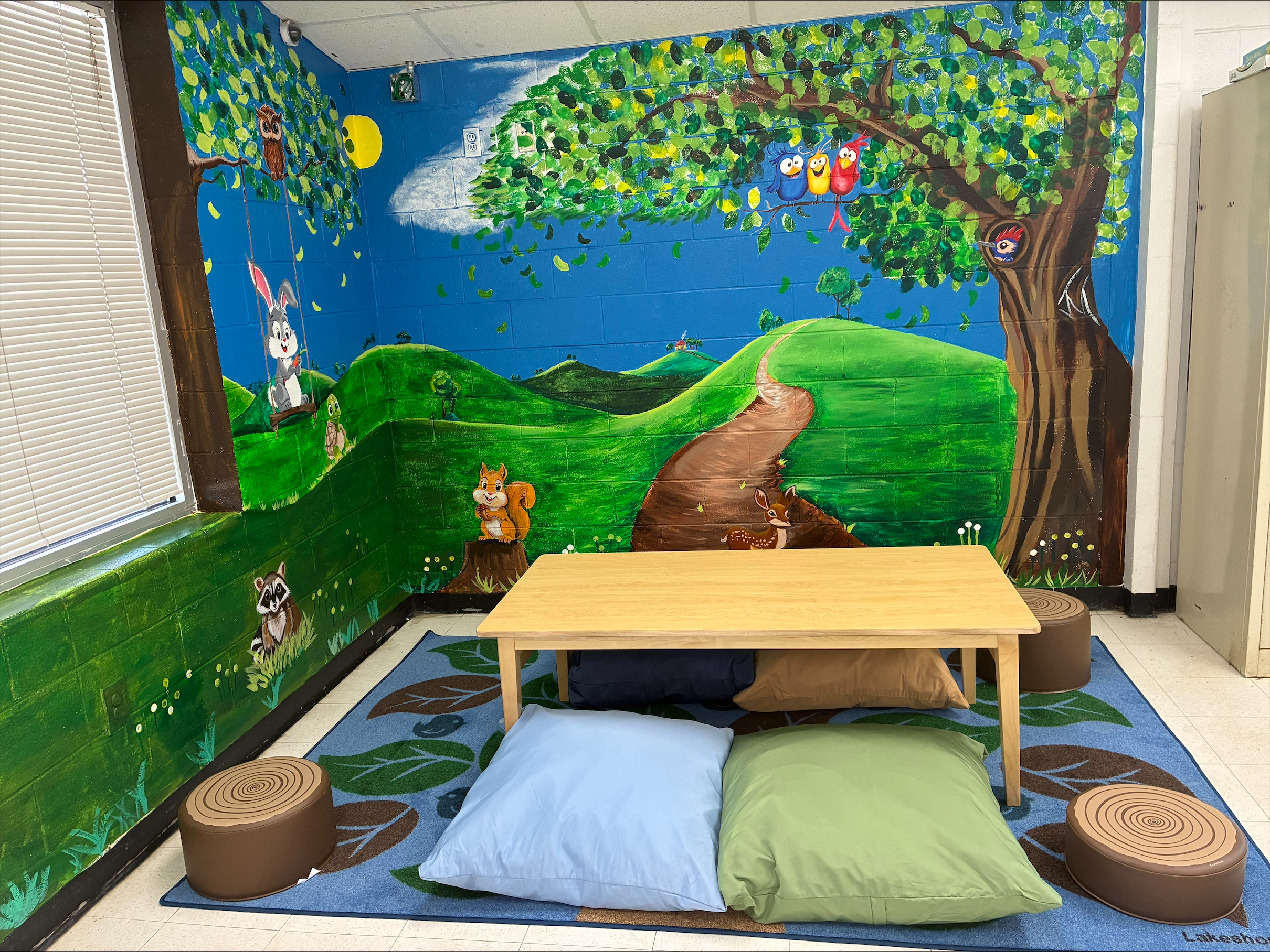 A colorful mural of a tree with birds and animals is painted on a prison wall. A small table and pillows sit in front of it to create a more child-friendly visitation space