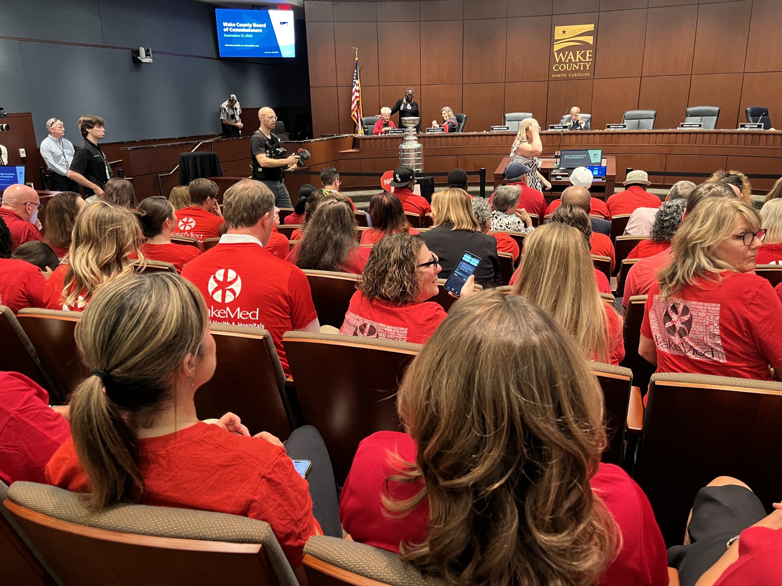 Shows the audience seats of a meeting room with at least six rows of people wearing red t-shirts with messages supporting WakeMed.