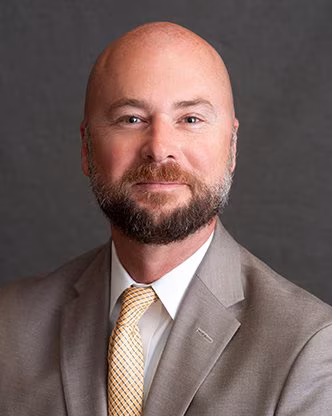 A white man with a bald head and brown beard wearing a tan suit with a yellow tie. He is John Bryant, vice president of operations for Pardee Hospital.