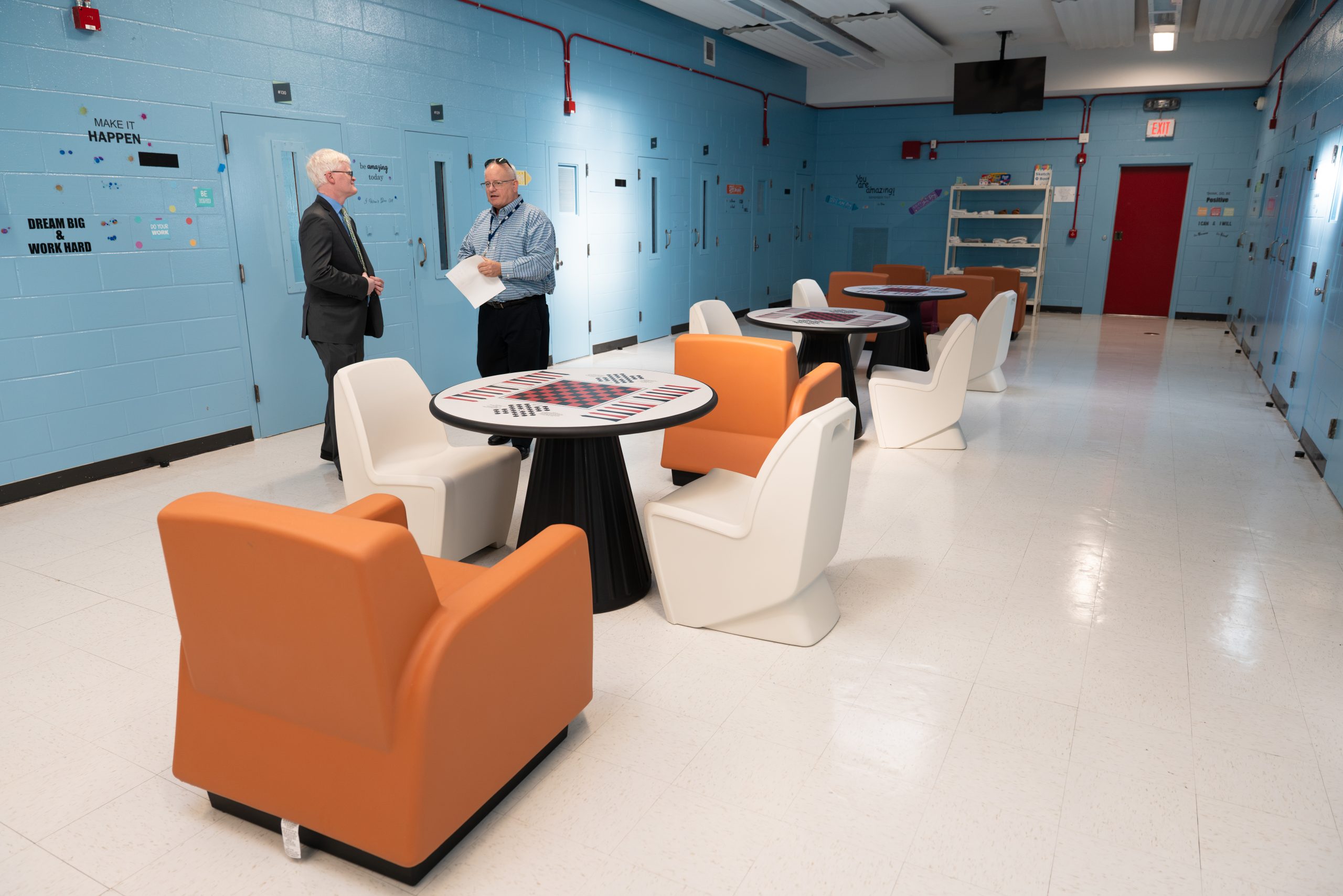 Two men stand in a juvenile justice mental health unit. The walls are painted bright blue and a line of tables are arranged in the open space