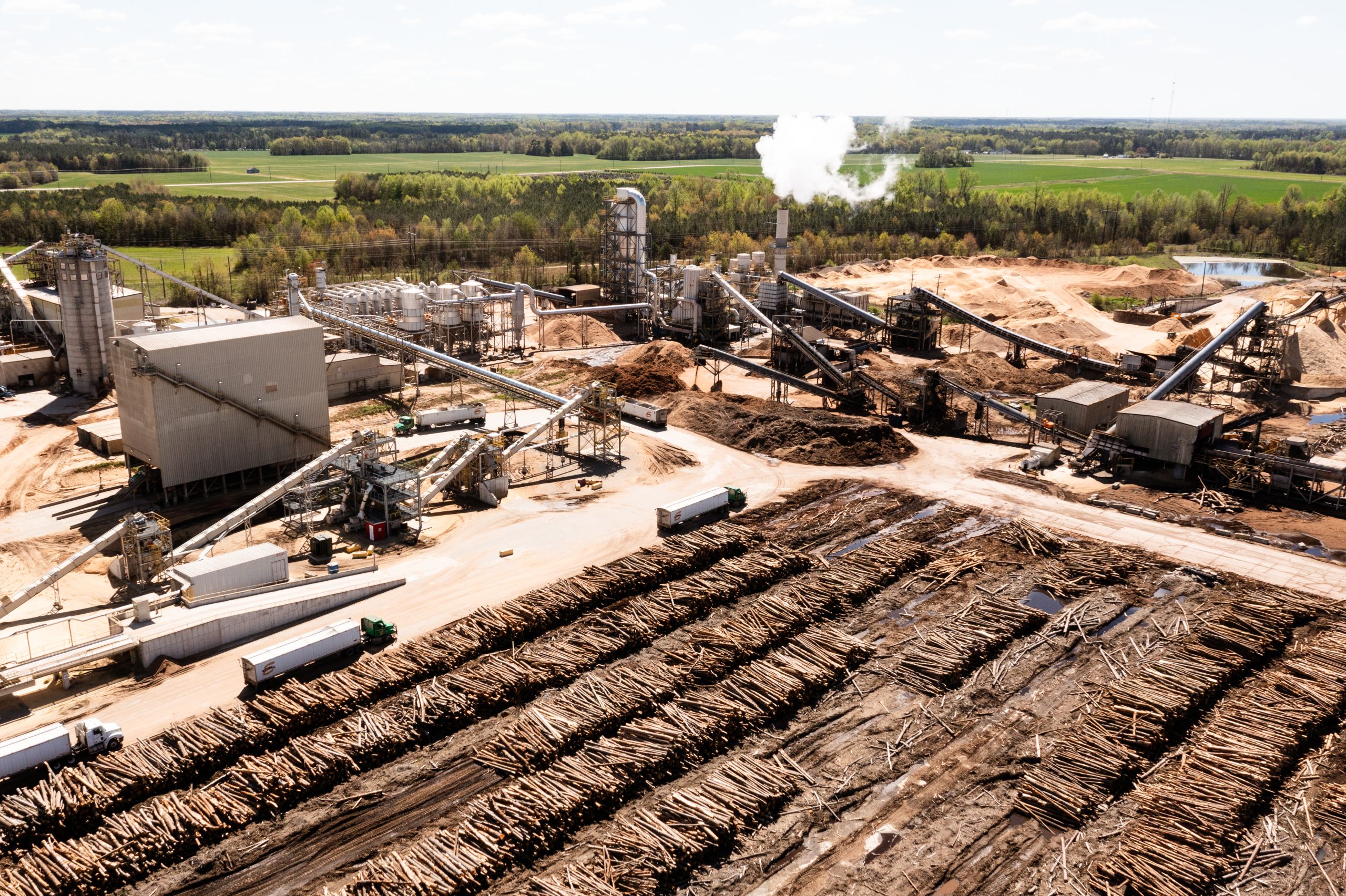 Aerial view of a wood pellet plant in Northampton County, with rows of stacked logs in the foreground, tractor-trailers on dirt roads, conveyors and large storage buildings, and a white plume rising from a stack. Farm fields and forest surround the site.