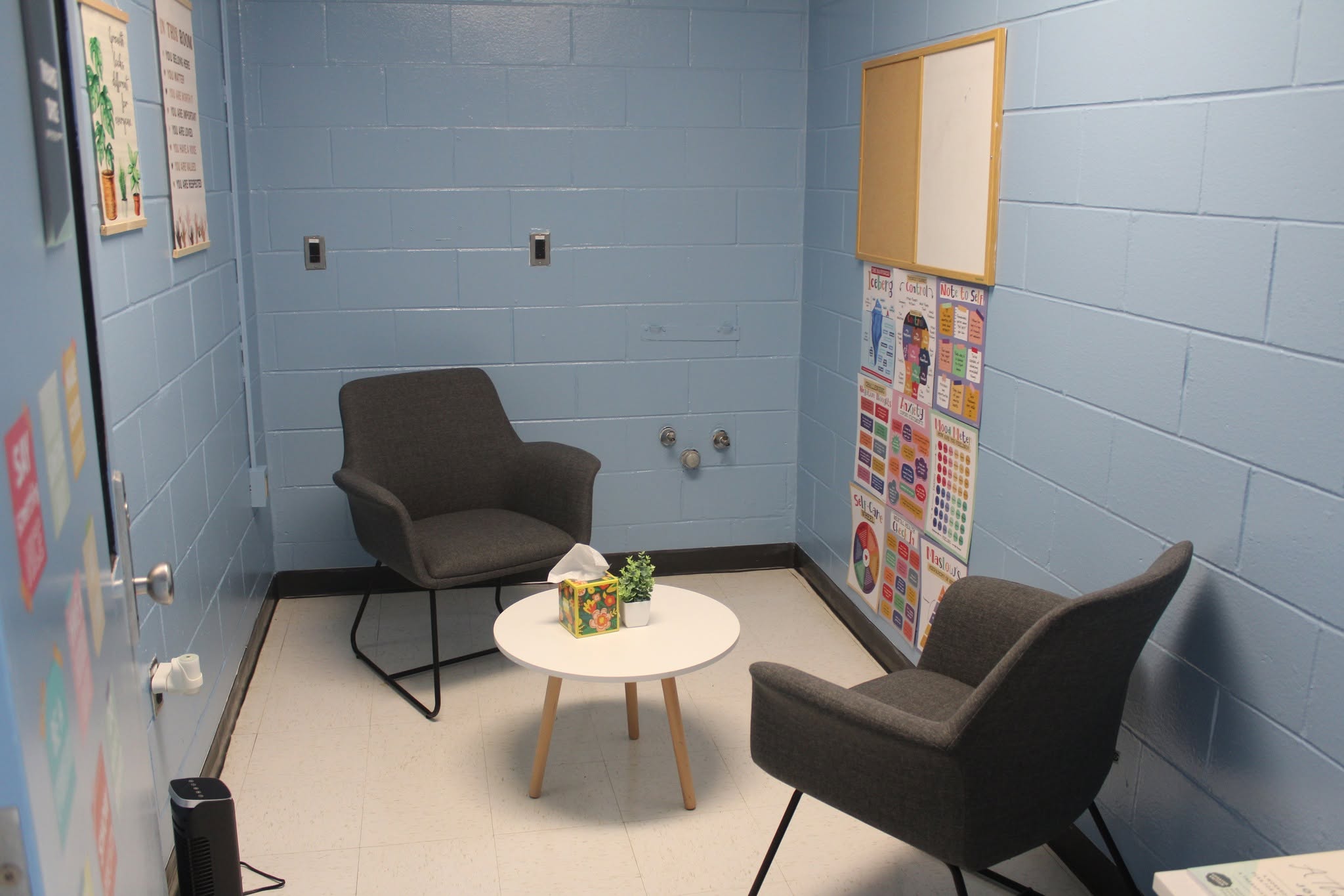 A room with cinder block wall painted bright blue with two gray chairs sitting around a small table where therapy sessions can happen in a specialized mental health unit inside a juvenile detention center.